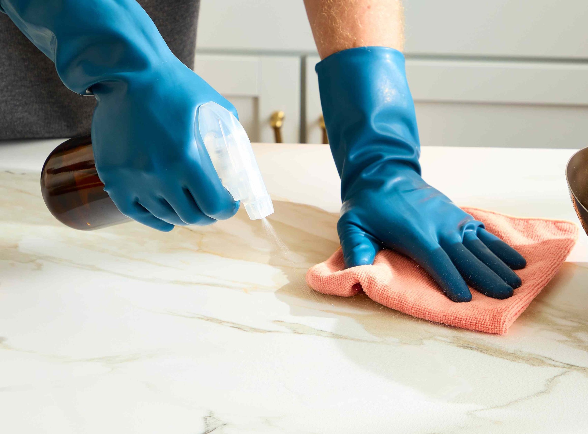 Professional cleaner in blue nitrile gloves applying eco-friendly disinfectant spray to a marble kitchen countertop—dusting, sanitizing, and polishing surfaces with pet-safe, non-toxic solution for deep home or apartment cleaning in Austin.