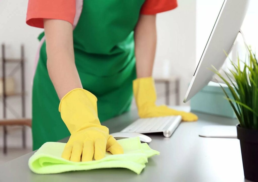 Professional cleaner in green apron and yellow gloves disinfecting office desk with eco-friendly microfiber cloth — same-day commercial cleaning service in Austin by Gleaming Space, pet-safe & baby-safe certified.