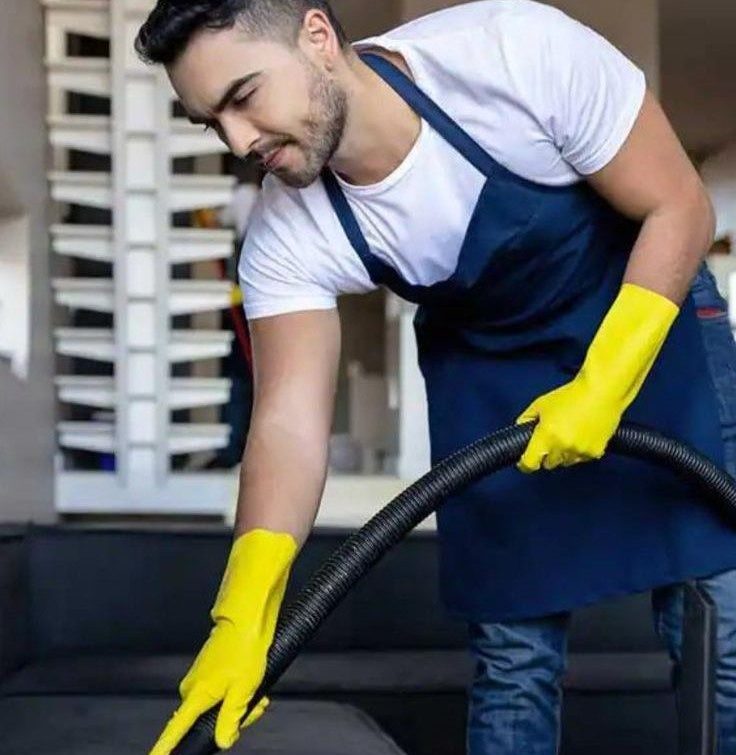 Professional cleaner in navy apron and yellow gloves using HEPA vacuum on sofa during one-time deep cleaning in Austin — eco-friendly, pet-safe home cleaning service by Gleaming Space.